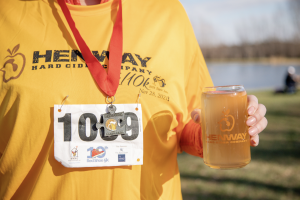A runner wearing their race shirt and race medal with a Henway Hard Cider in hand. 