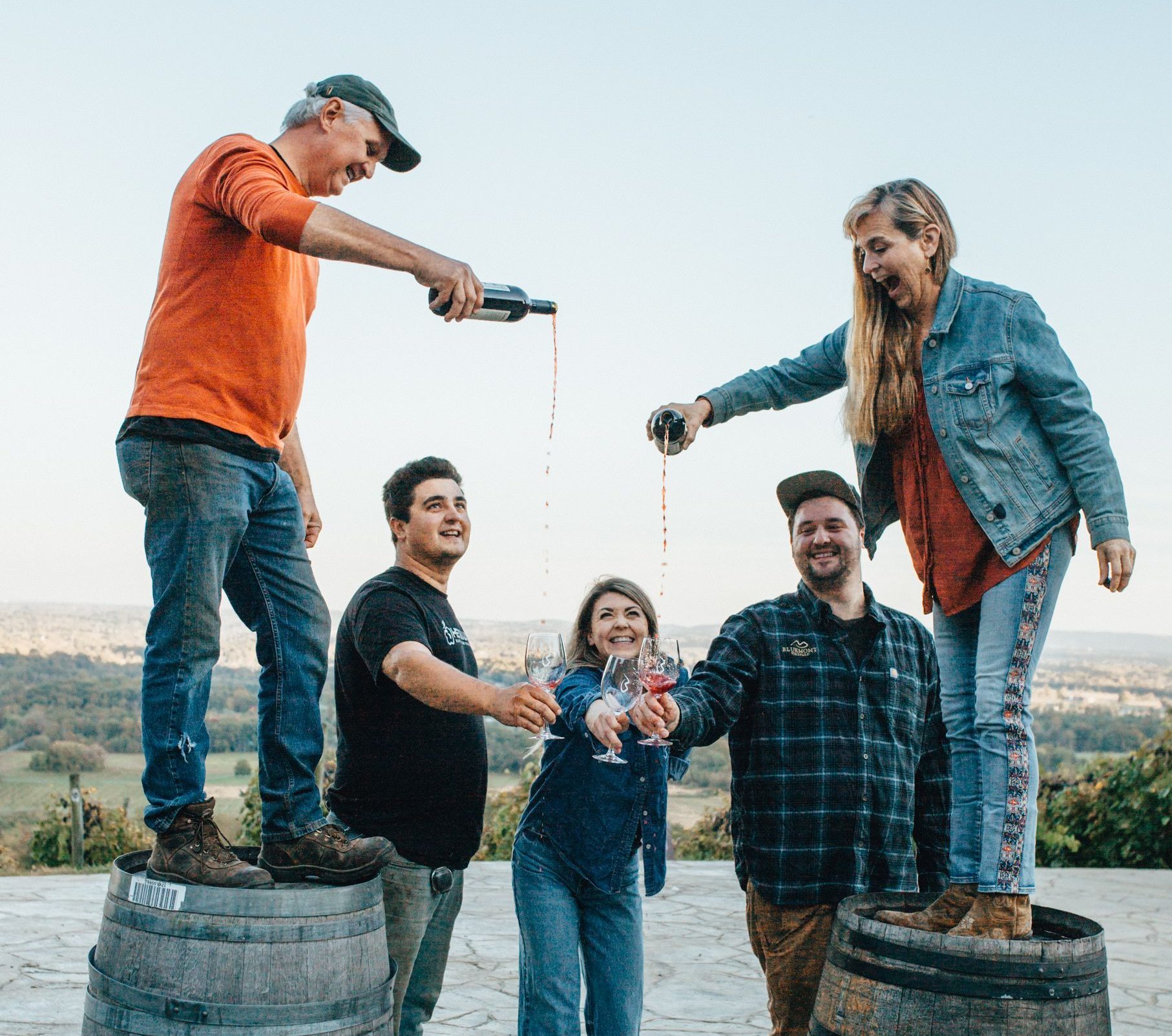 The two Farmers and Founders standing on wine barrels pouring wine while three others stand below holding out their wine glasses to be filled