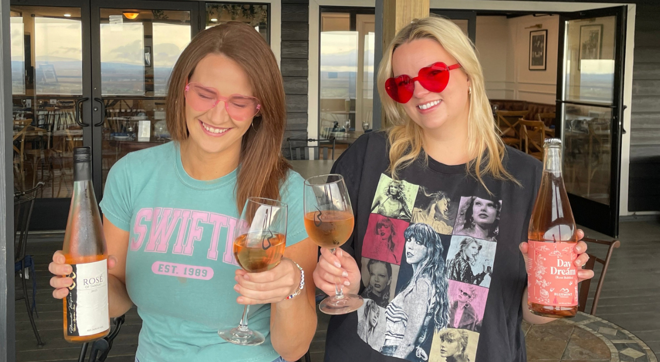 Swiftie Brunch main image. Two girls, each holder a bottle of rose in one hand and a glass of rose in the other. Both wearing Taylor Swift t-shirts