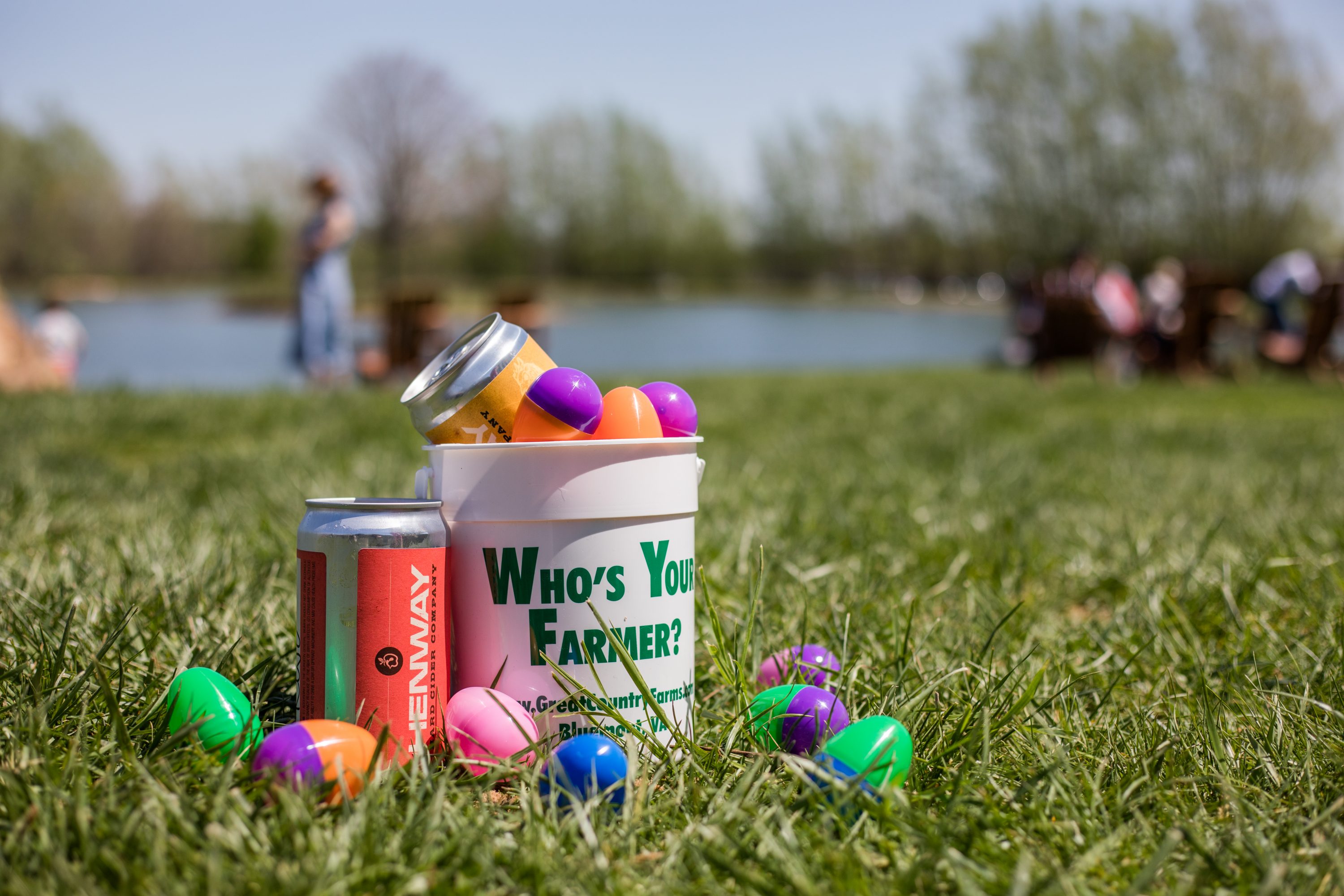 Adult Egg Hunt photo of a Great Country Farms egg bucket filled with Easter eggs and a Henway Hard Cider crowler with eggs scattered around the base of the bucket in the grass